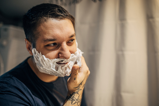 A young handsome man is standing in front of a bathroom mirror and applying shaving cream to his face