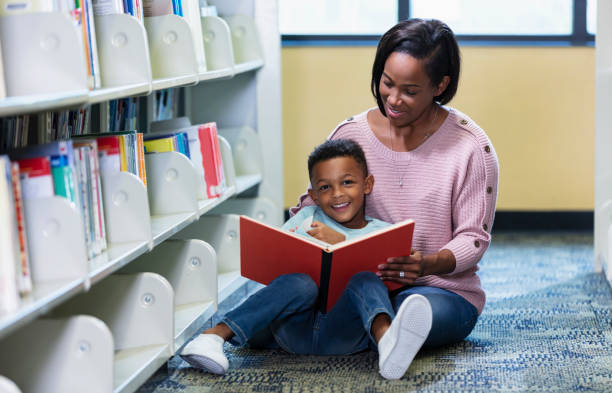 An African-American mother and son reading together in the library. The 5 year old boy is sitting on his mother's lap, on the floor by a bookshelf. He is smiling at the camera.