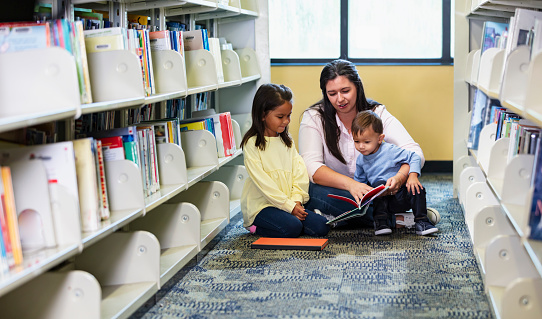 A young family sitting on the floor of a library between bookshelves, reading a book. The son, a toddler, is sitting on mother's lap, and the 5 year old daughter is next to them, listening as mother reads aloud. The children are multiracial, Asian and Caucasian.