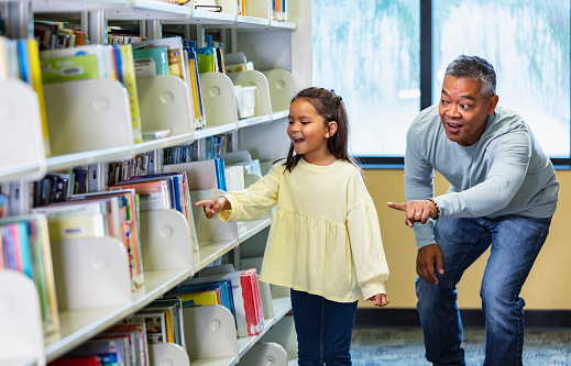 An Asian father and his multiracial daughter looking for a book together in the library. They are standing next to a bookshelf, and dad is bending down to her eye level. They both look excited, pointing to a book on the shelf.