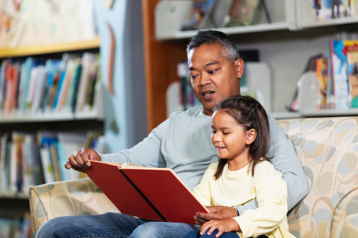 An Asian father and multiracial daughter reading together in the library. The 5 year old girl is sitting next to her father on a comfortable armchair by a bookshelf, looking at the book as he reads to her.