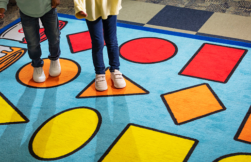 Cropped view of two 5 year old children playing on a colorful rug in a playroom, standing on different shapes.