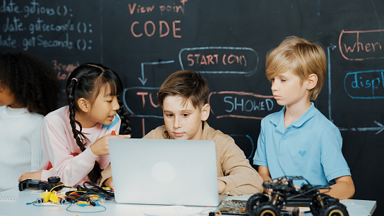 Closeup of boy using laptop programing engineering code and writing program while group of smart diverse student standing surrounded by friend in STEM technology classroom at blackboard. Erudition.