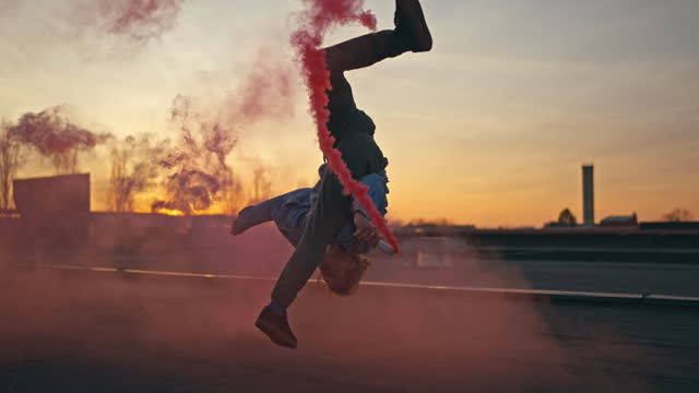 SLO MO Active Young Man Performing Breakdance while Holding Smoke Bombs on Rooftop at Sunset