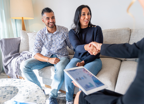 Mature couple meeting with financial advisor or real estate agent at home. They are sitting on a sofa and shaking hands looking at a digital tablet presentation.