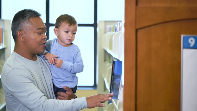 Asian father and toddler in library looking at bookshelf