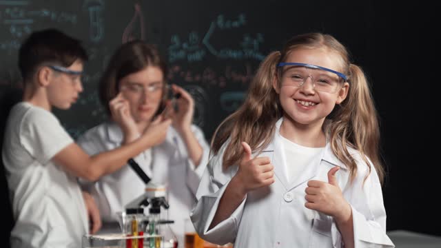 Caucasian girl showing thumb while teacher doing experiment behind. Erudition.