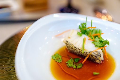 Chinese-style steamed cod fish with soy sauce is beautifully presented on a white plate in a restaurant, reflecting the elegance and deliciousness of this culinary creation.