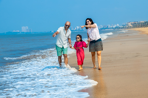 Happy indian Family of three walking on tropical beach and enjoying summer vacation. mother holding hands of her daughter point aside. friendly family Concept.