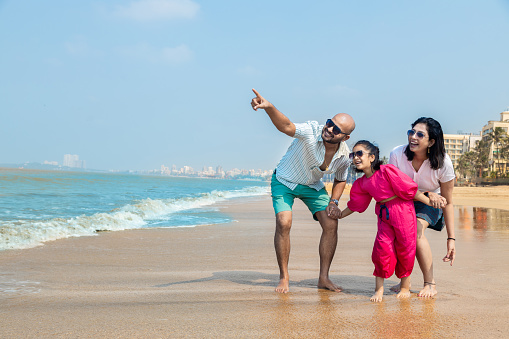 Happy indian Family of three enjoying summer vacation on tropical beach. father point aside with mother and daughter looking. friendly family Concept.