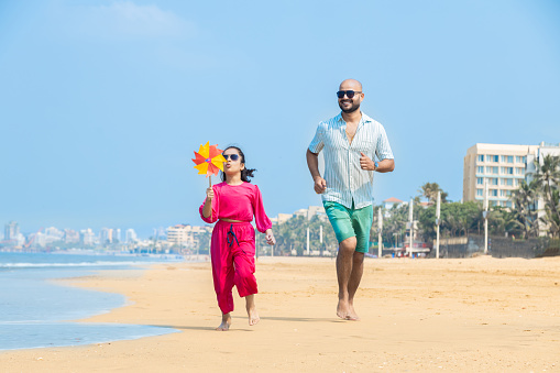 Indian father and daughter running on tropical beach and enjoying summer vacation. daughter playing with paper windmill on sea side.
