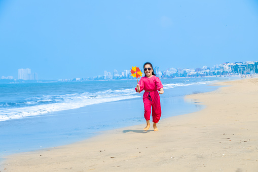 Beautiful young indian girl playing with paper windmill on tropical beach and enjoying summer vacation.