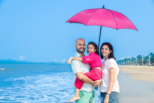 Happy indian family of three standing under sun umbrella enjoying summer vacation on tropical beach. Father holding her daughter on beach.
