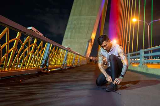 Asian man athlete in sportswear tie shoelaces during jogging exercise in the city at night. Healthy man enjoy outdoor lifestyle do sport training running fitness workout through the city street at night.
