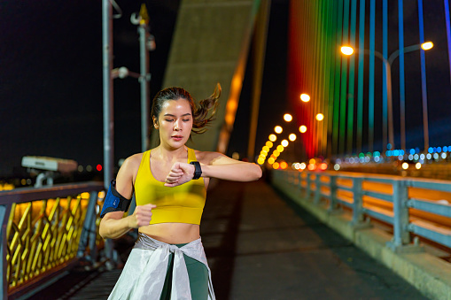 Asian woman athlete in sportswear using smart watch tracking fitness goals and checking cardio heart rate during jogging exercise in the city at night. Attractive girl enjoy outdoor lifestyle do sport training running workout at night.