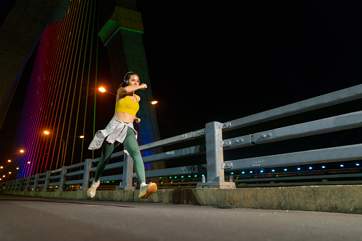 Asian woman athlete in sportswear listening to the music on headphones and smartphone during jogging exercise in the city at night. People enjoy outdoor lifestyle do sport training running fitness workout through the city street at night.