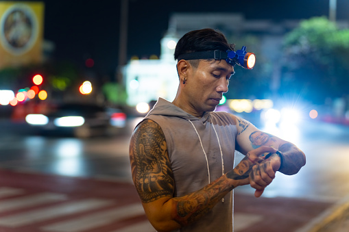 Asian man athlete in sportswear and headlamp using smart watch tracking fitness goals checking cardio heart rate during jogging exercise in the city at night. Sportsman body builder enjoy outdoor lifestyle do sport training running workout at night.