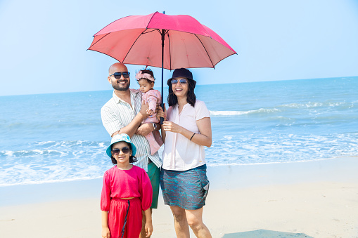 Happy indian family of four standing under sun umbrella enjoying summer vacation on tropical beach. father holding her little daughter on beach Happy Family Concept.