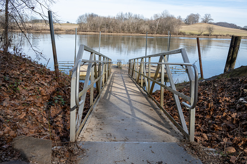 A dock leading to a lake in the winter with brown leaves and a hand rail.