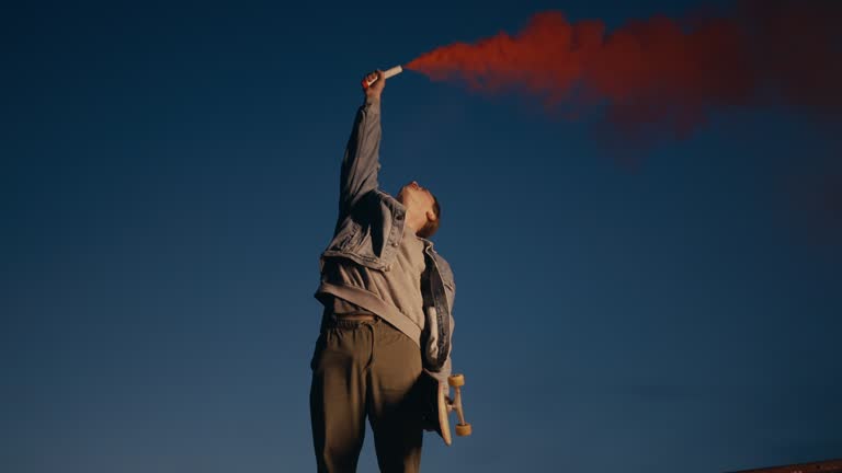 SLO MO Young Male Skateboarder Raising Arm While Holding Smoke Bomb with Fume on Rooftop at Night