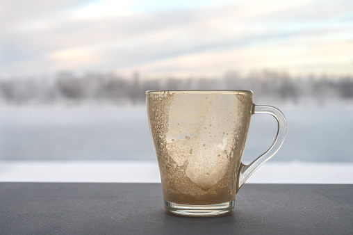 An empty glass cup of cappuccino on a table against the backdrop of a window with a blurred winter landscape.