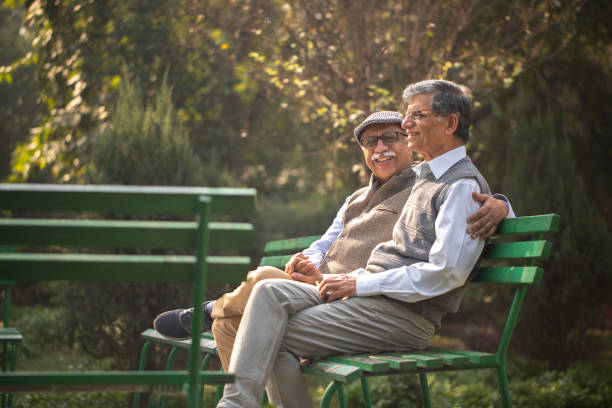 Retired senior male friends dressed in warm clothes smiling and talking while sitting on bench in park
