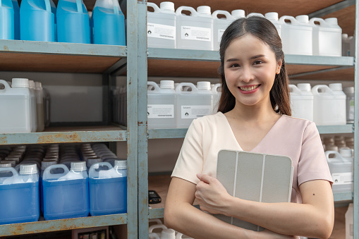 Beautiful Asian female employee is checking chemical products (solvent) on the shelves with a digital tablet.