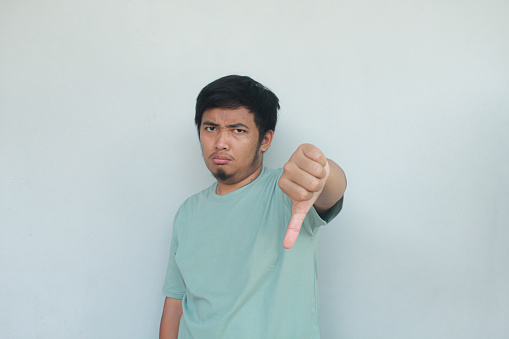 A young Asian man in a green t-shirt  looking unhappy and angry showing rejection and negative with thumbs down gesture. Bad expression. Isolated in gray background.