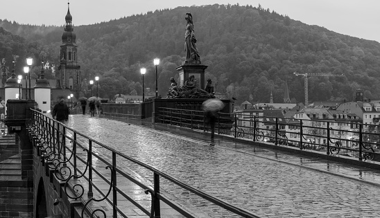 Black and white photo of the old bridge (Karl-Theodor-Brücke) in Heidelberg during a rainy day. Some pedestrians are walking with umbrellas. Blurred movement effect of the people on the bridge.