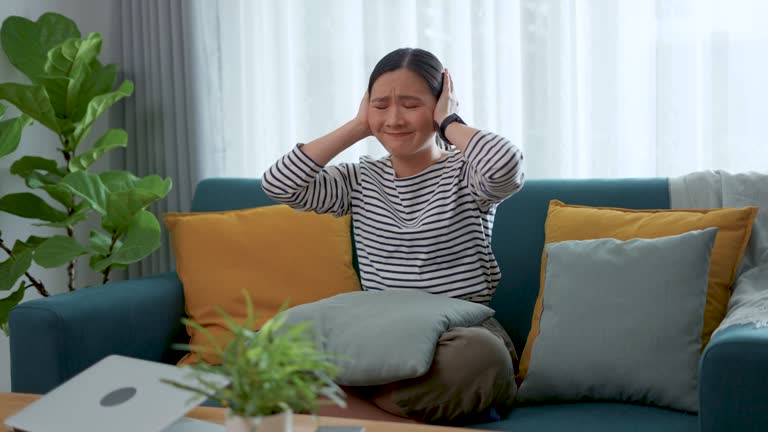 Asian woman feel annoyed noise covering ears with hands sitting at home.