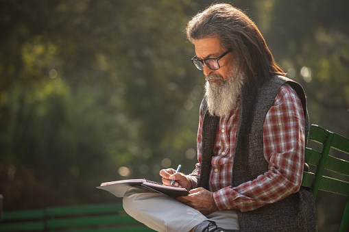 Senior man with beard and long hair thoughtfully and writing in book while relaxing on bench in park