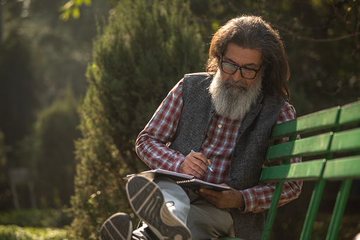 Senior man with beard and long hair looking away thoughtfully and writing in book while relaxing on bench in park
