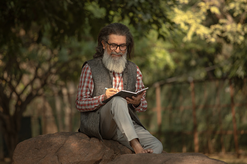 Full length of focused bearded senior man writing in diary while sitting on rock against trees in garden