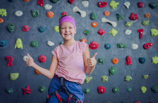 A young girl in sporty attire is climbing an indoor rock wall, wearing a safety harness. She grips the colorful climbing holds while looking back with determination. The well-lit space and professional setup make this an ideal environment for developing strength and confidence.