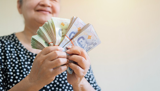 Happy older Asian woman holding a lot of Thai money that she has won, feeling excited. Retirement concept.