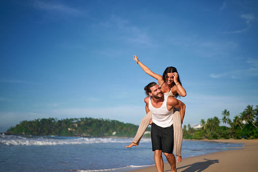 Man carries woman on his back, both smiling. Joyful couple enjoys piggyback ride on sunny beach. Sea waves, clear sky, casual summer attire. Holiday, romance, travel, fun moments by ocean.