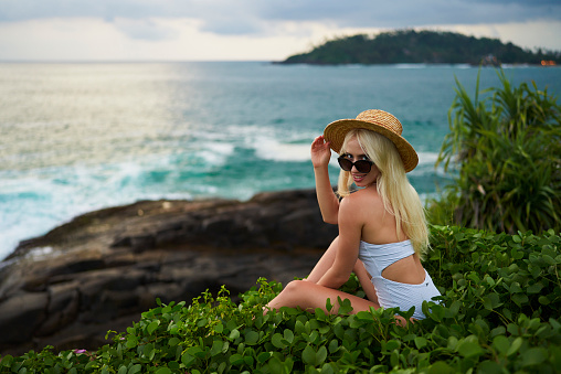Blonde tourist relaxes by seaside, smiles towards horizon. White bikini-clad traveler amidst greenery on a rocky cliff, represents holiday getaway, serenity, and exploring exotic locations.