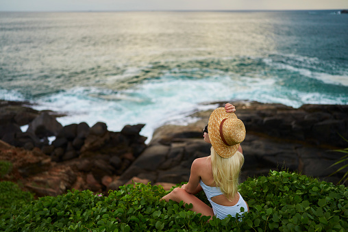 Female adventurer in summer attire sits peacefully atop a seaside bluff, overlooking the turbulent waves, capturing the essence of solitude and reflection for lifestyle and tourism imagery.