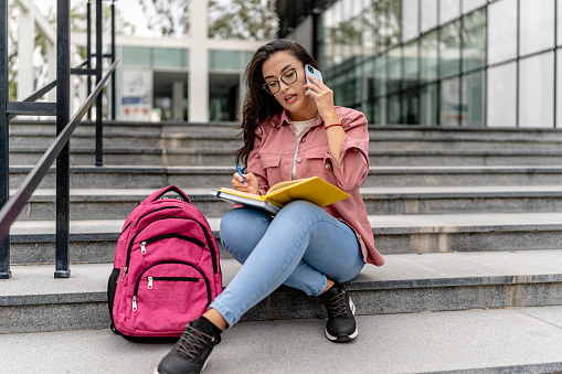 A female student is captured in a candid moment, quickly checking her phone while holding a notebook, ready to enter her university building