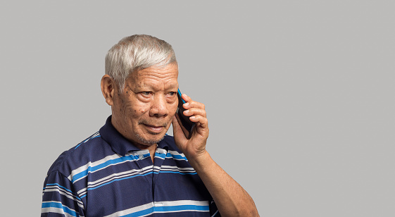 Happy Asian senior man using a smartphone while standing on a gray background. Aged people and communication concept