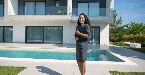 Happy successful female real estate agent offering key, showing bunch at camera, posing for professional real estate agent portrait with rental accommodation and couple of renters, buyers in blurred background