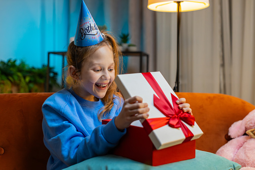 Happy Caucasian child girl wears festive birthday cap hat hold gift box with ribbon congratulating. Teenager female kid celebrating party event, opening delivery greeting present at home room on sofa