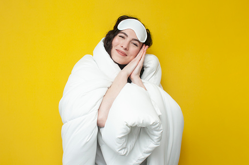 cute young girl in sleep mask and with warm soft comfortable blanket stands on yellow background, the woman is ready for bed, the concept of healthy sleep