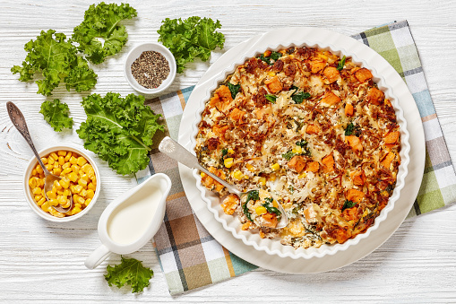 Sausage-Hash Brown Breakfast Casserole with corn, kale, sweet potatoes and cheese in baking dish on white wooden table, horizontal view from above