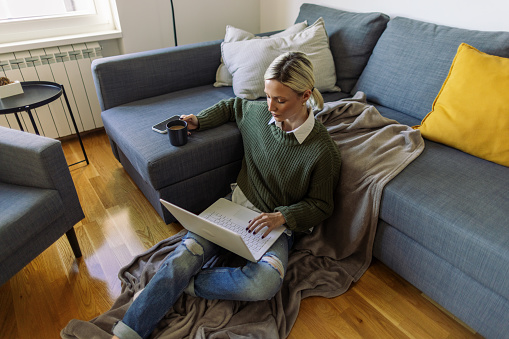 A young woman sitting on the floor of her living room, typing on her laptop and drinking tea or coffee. Mobile phone is next to her.