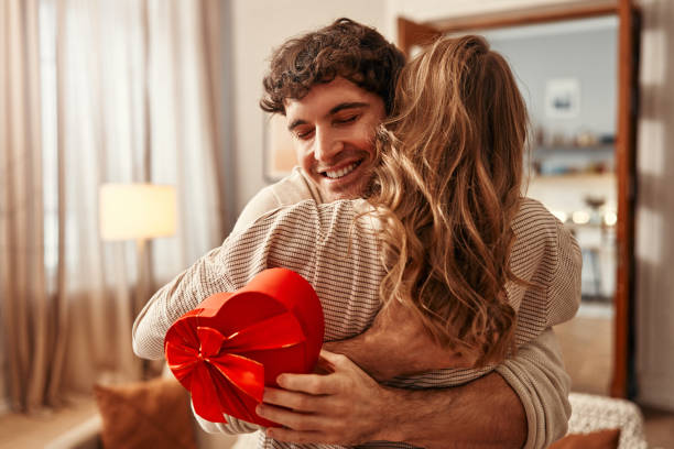 Valentine's Day. Couple in love at home. Happy Valentine's Day. A man gives a heart-shaped gift box to his beloved woman in the living room at home, the woman hugging him tenderly. Romantic evening together. gift for girlfriend stock pictures, royalty-free photos & images