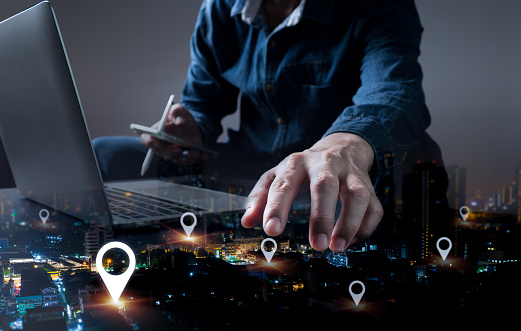 Mark the location of the address on a GPS map or mobile app. A man is using a laptop and a tablet to look for a certain address in a nocturnal metropolis; his hand is partially visible.