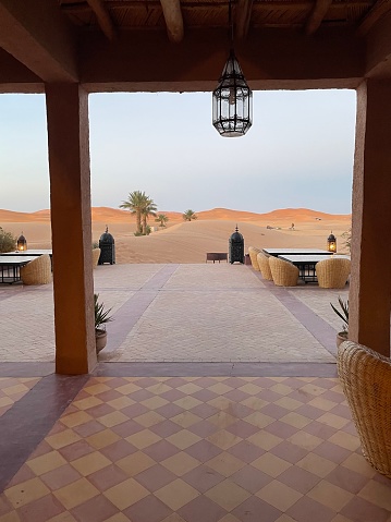 Large terrace with views of the sand dunes of the Sahara Desert, and palm trees. Square tiled floor, square tables and outdoor armchairs, light fixtures. Clear sky