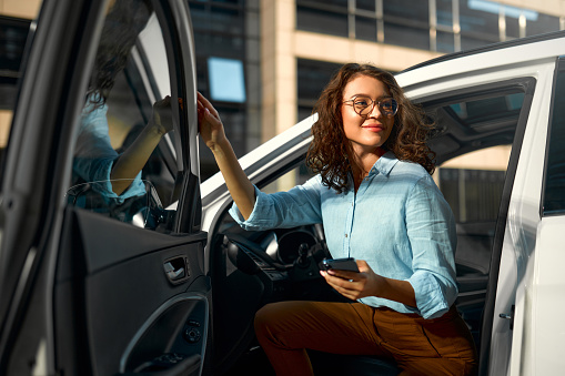 Beautiful young happy caucasian business woman sitting in her new car and using smartphone. Buying and renting a car. Travel, tourism, recreation.