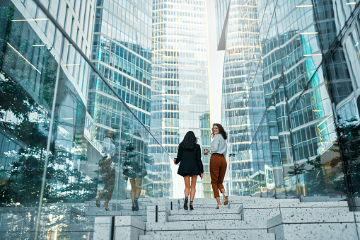 A team of successful business women of different races is walking up the steps, discussing strategy, drinking coffee. Various women walk along the street against the backdrop of buildings.